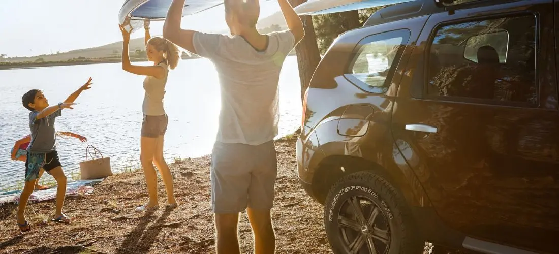 Eine Familie lädt ein Surfbrett auf das Dach eines dunklen SUVs an einem sonnigen See