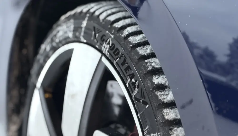 En närbild av ett Goodyear Vector All Season 4-däck på en mörkblå bil, där mönsterblocken och sidoväggen är täckta av nysnö bredvid en silverfärgad fälg med flera ekrar.