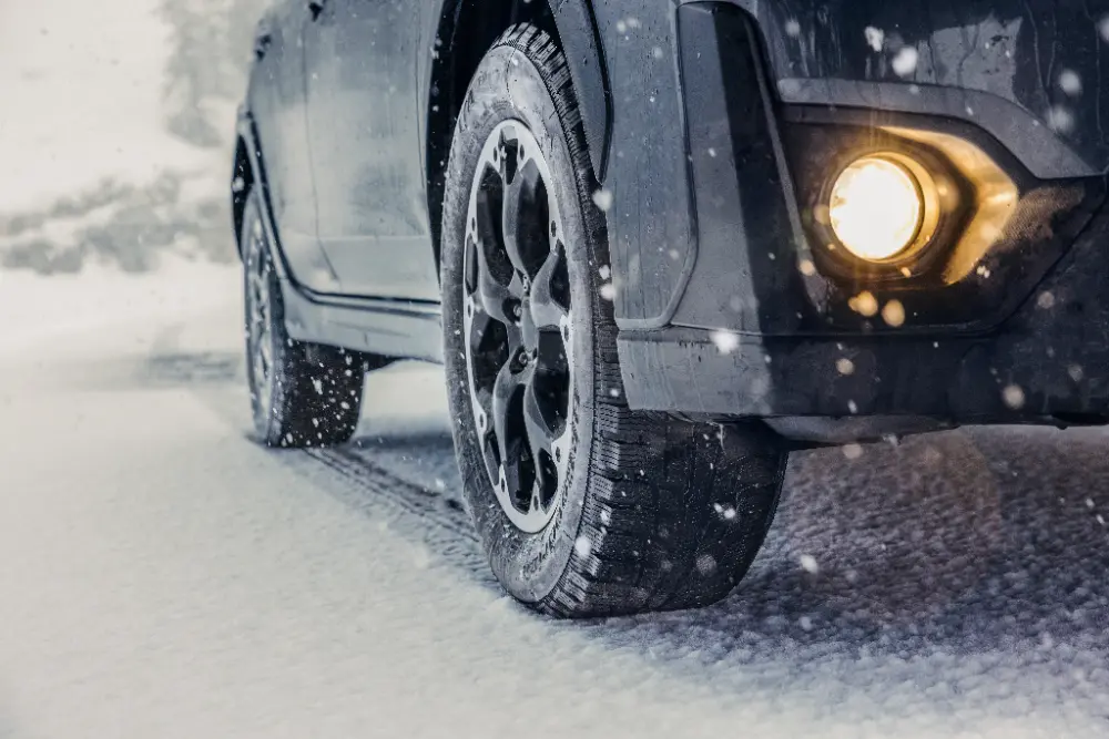 Primer plano de la rueda delantera y el faro de un todoterreno oscuro que circula bajo la nieve que cae en una carretera invernal.