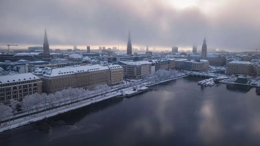 Luftaufnahme der Hamburger Innenstadt im Winter, mit schneebedeckten Dächern und von Bäumen gesäumten Promenaden entlang der Binnenalster