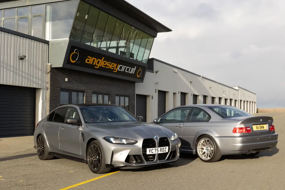 BMW M3 Competition grigia, parcheggiata accanto a una BMW M3 CSL E46 argento, fotografata davanti all’Anglesey Circuit in vista di un confronto diretto.