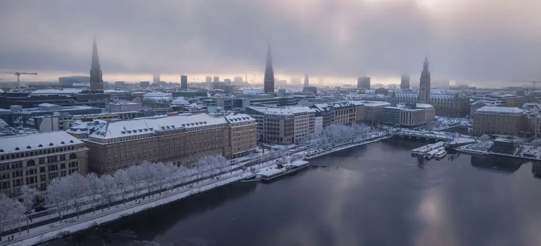 Luftaufnahme der Hamburger Innenstadt im Winter, mit schneebedeckten Dächern und von Bäumen gesäumten Promenaden entlang der Binnenalster