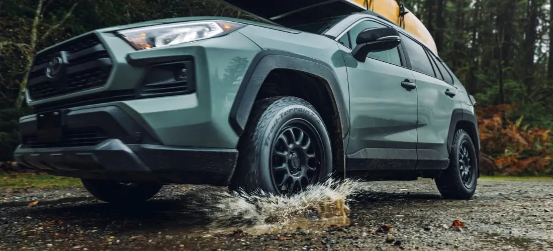Die Frontpartie eines grünen Geländewagens durchquert eine Pfütze auf einem Schotterweg, Wasser spritzt unter dem BFGoodrich-Reifen hervor
