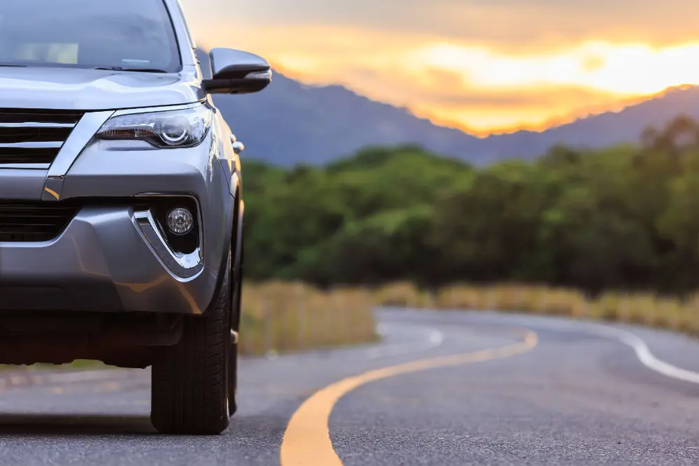 Silverfärgad SUV parkerad på en slingrande bergsväg i solnedgång, med strålkastare och frontgrill synliga.
