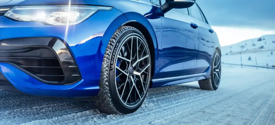 Die Nahaufnahme zeigt Michelin CrossClimate und Frontstoßstange eines blauen Fahrzeugs auf einer schneebedeckten Straße