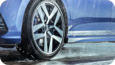 Primer plano de un neumático y un vehículo azul circulando por una carretera mojada